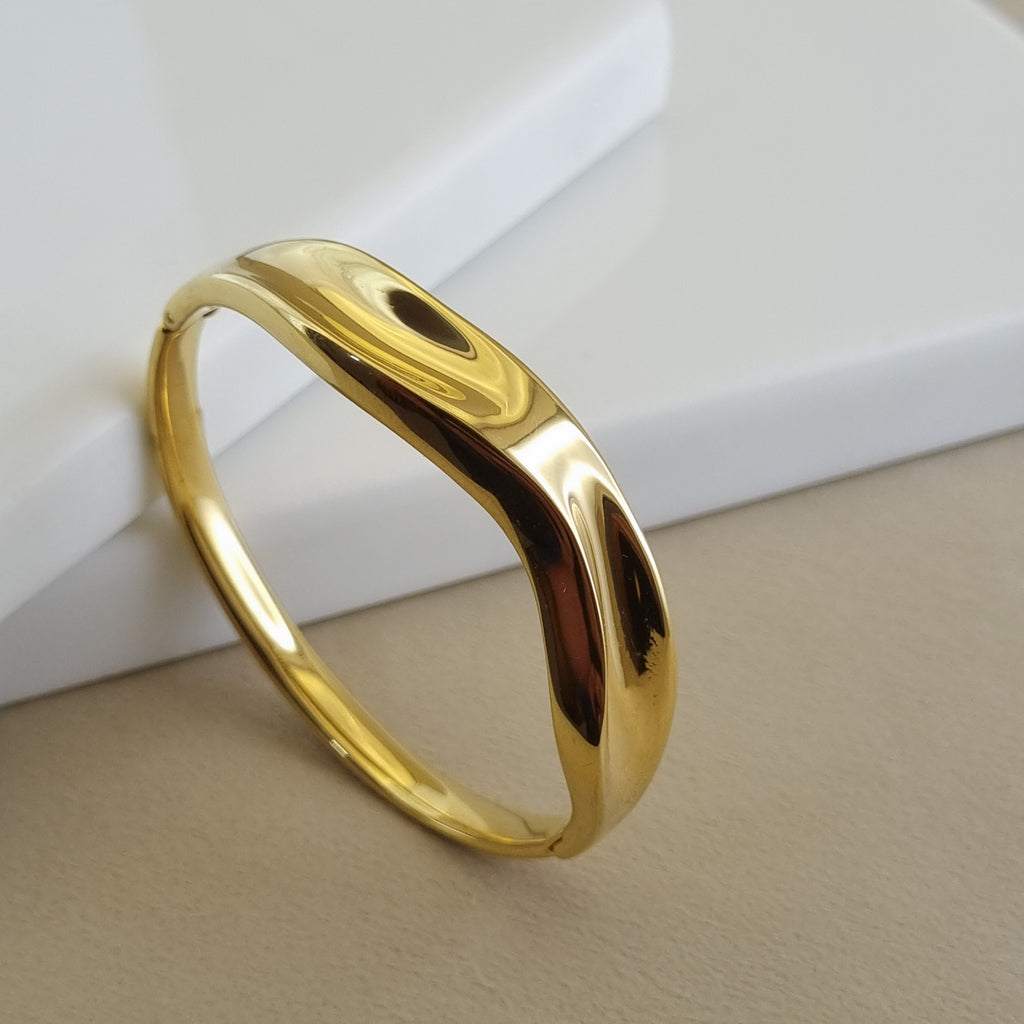 A high-polish 18K gold plated organic wave bangle with a molten texture, shown resting on a minimalist white and beige background.