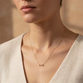 A woman wearing a delicate 18K gold-plated necklace featuring a central crystal-encrusted infinity symbol pendant on a minimalist beige background.