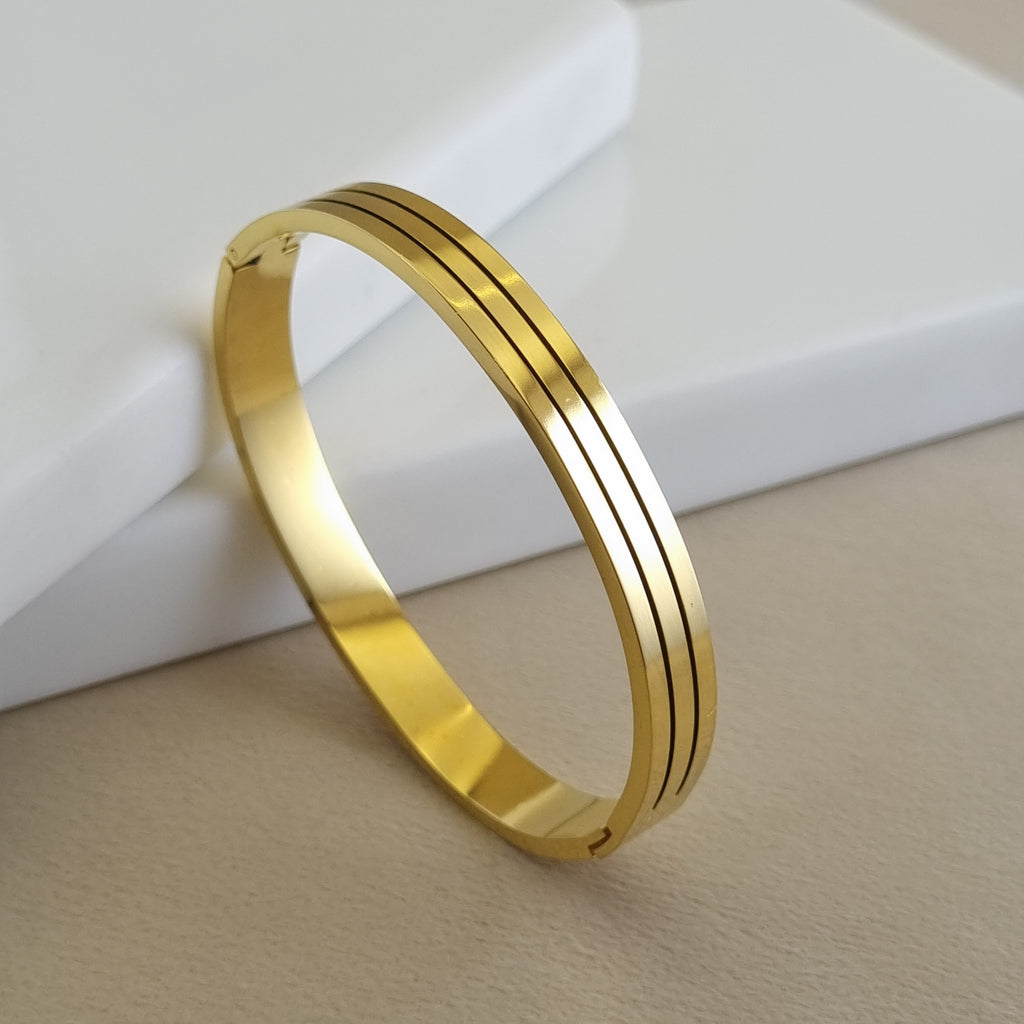 A side-view of a Treshire 18K gold-plated bangle featuring three parallel vertical grooves, displayed on a neutral, minimalist surface.