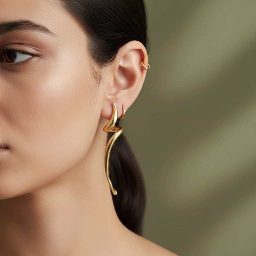 A woman with a sleek ponytail wearing large, 18K gold-plated sculptural earrings in a fluid, molten flame-like design that climbs the earlobe.