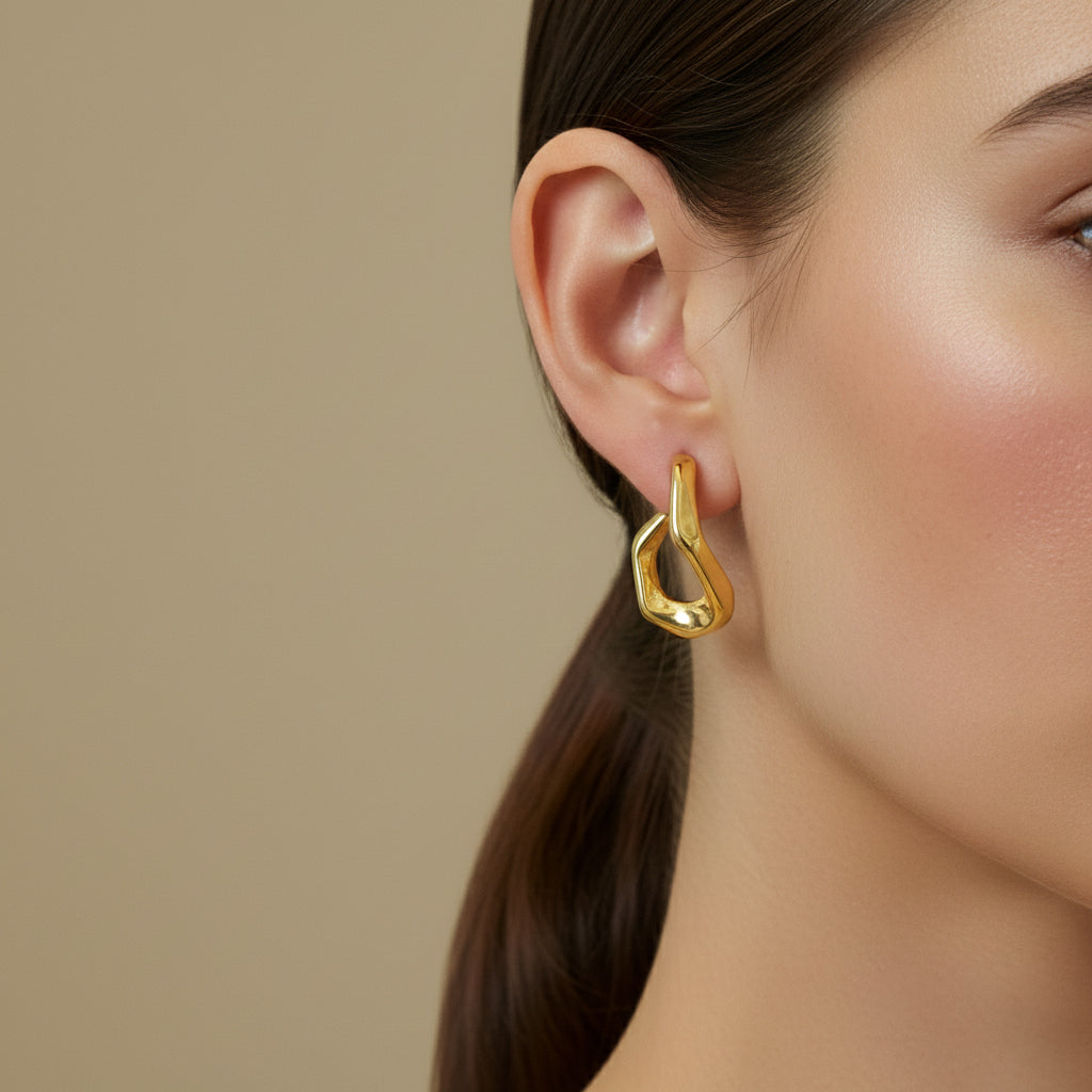 A woman with a sleek low ponytail wearing 18K gold-plated asymmetrical geometric huggie earrings with a high-polish reflective finish.