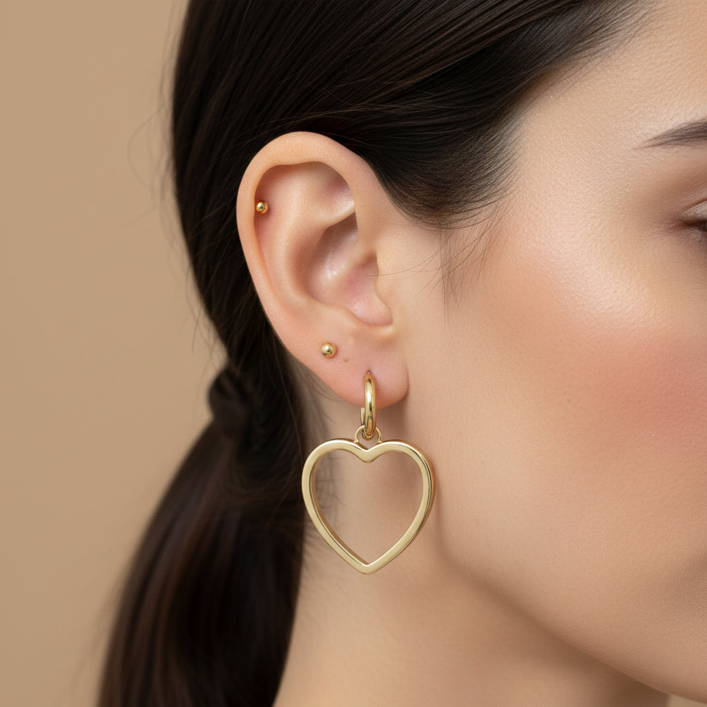 A woman with a sleek ponytail wearing 18K gold-plated huggie hoop earrings with a large open-heart shaped drop and a high-polish finish.