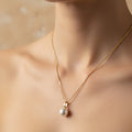 A woman wearing a delicate 18K gold-plated necklace featuring a single, high-luster round pearl pendant on a minimalist background.