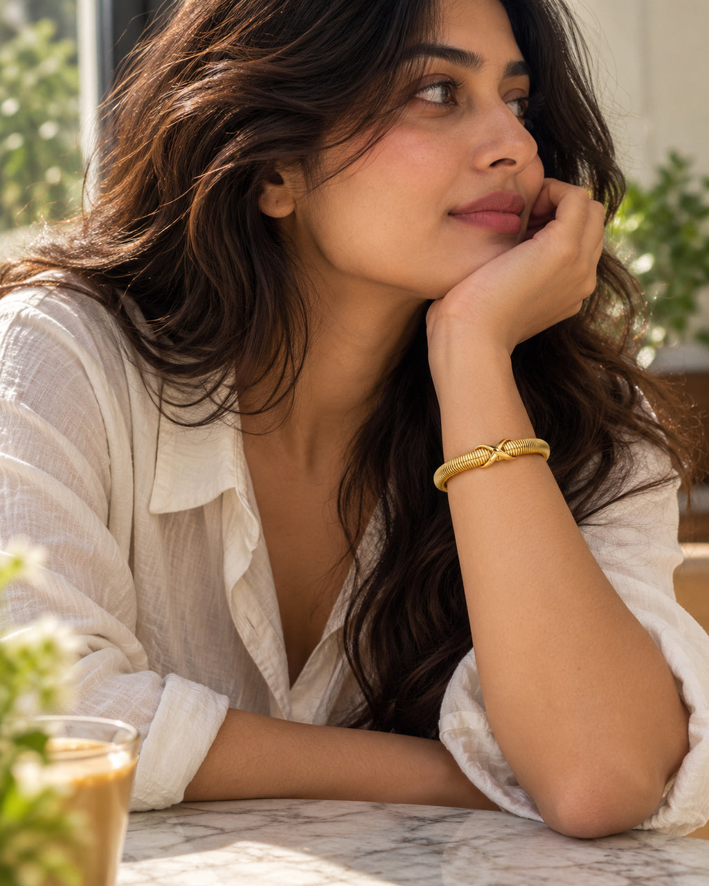 A woman in a white blouse rests her chin on her hand, wearing a gold bangle bracelet with a small knot detail, in a softly lit indoor setting.
