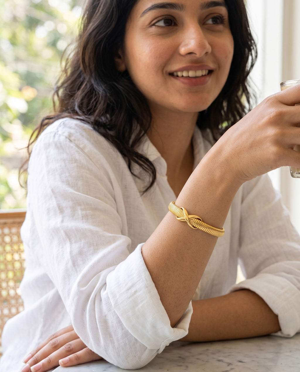 A woman wearing a gold knot bracelet on her wrist, posing in a white linen shirt at a table.