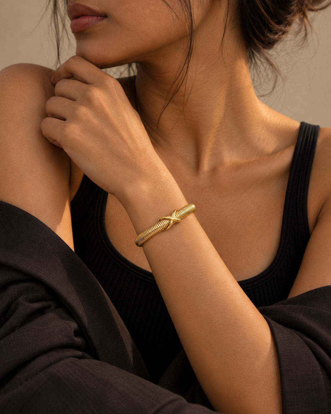 A close-up lifestyle shot of a model wearing a textured gold bangle bracelet with a star detail, paired with a black ribbed tank top and dark draped fabric.