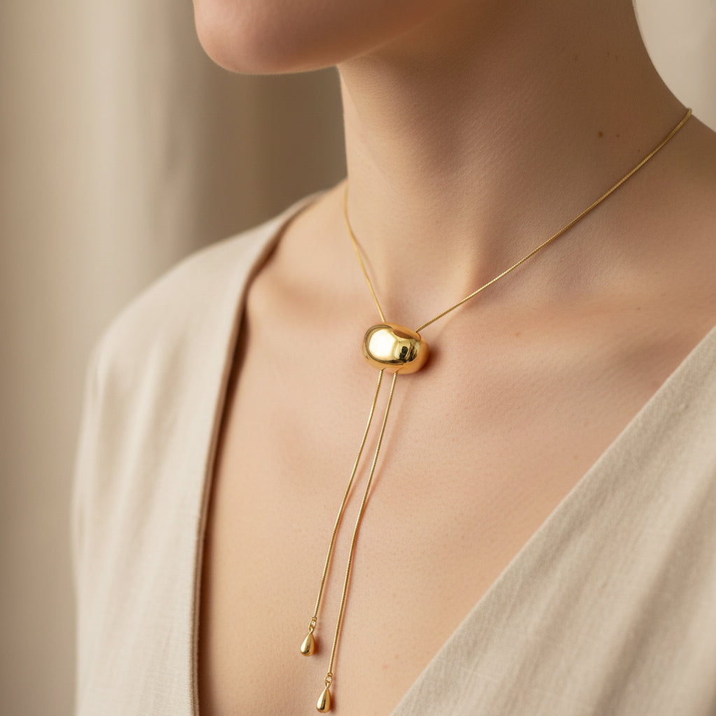 A woman wearing an 18K gold-plated lariat necklace with an adjustable molten-style orb slide and weighted gold droplets on a minimalist beige background.