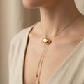 A woman wearing an 18K gold-plated lariat necklace with an adjustable molten-style orb slide and weighted gold droplets on a minimalist beige background.