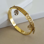 A high-polish 18K gold-plated triple-band bangle featuring five circular Tree of Life charms, displayed on a minimalist white and tan surface.