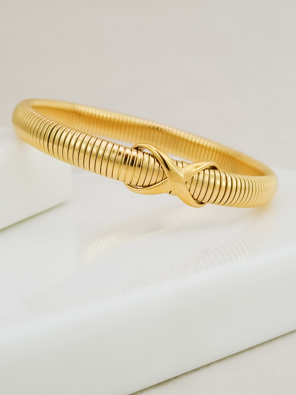 A gold-colored textured snake-style bangle bracelet with an infinity symbol at the center, resting on a white surface.