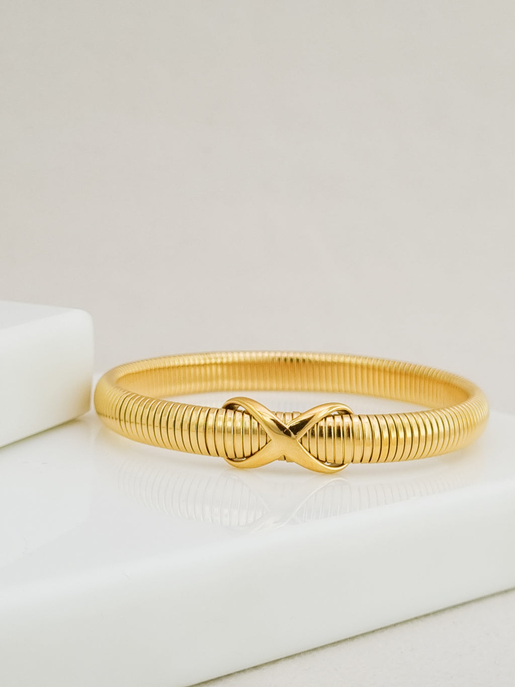 A gold-colored snake-style bangle bracelet with a central infinity symbol clasp, resting on a white surface.