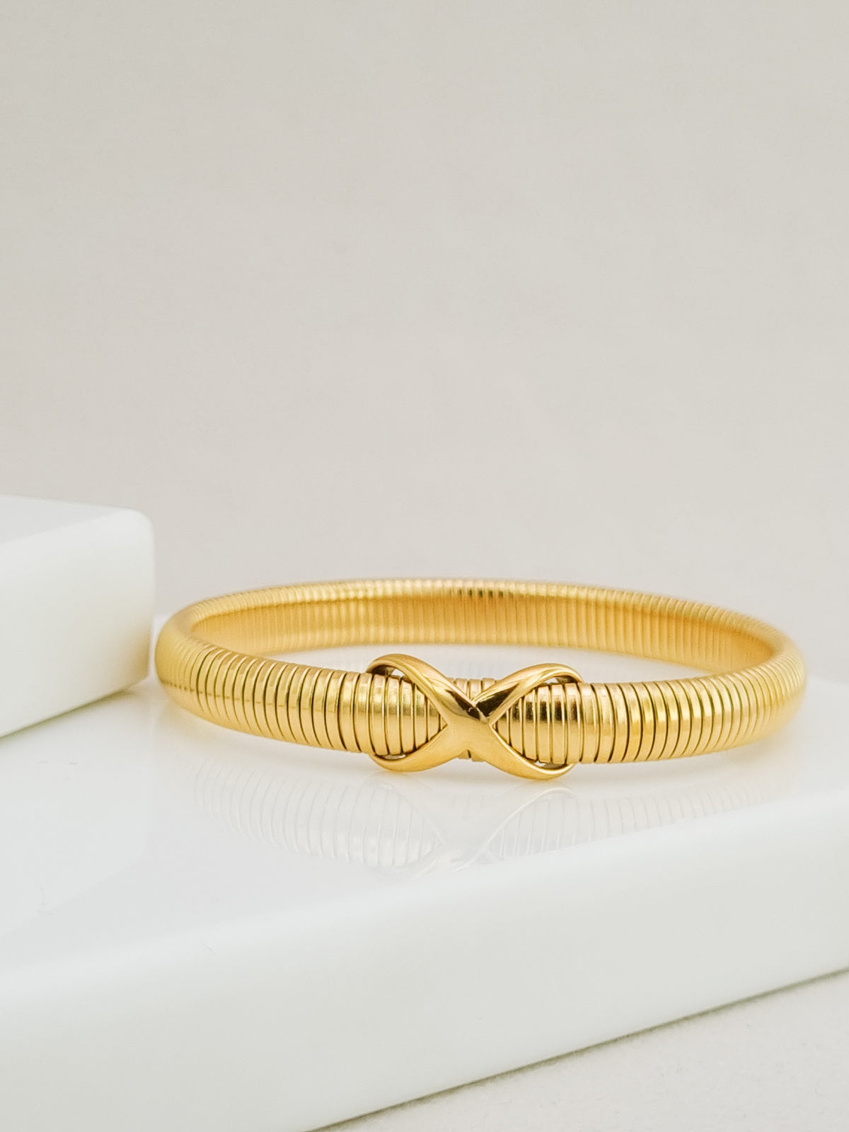 A gold-colored snake-style bangle bracelet with a central infinity symbol clasp, resting on a white surface.