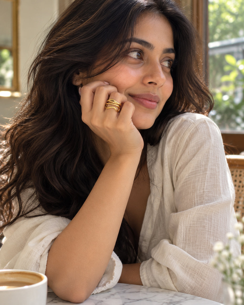 A woman with long dark wavy hair rests her chin on her hand, wearing a white textured blouse and gold rings, in a softly lit indoor setting.