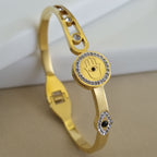 An 18K gold-plated bangle featuring a crystal-encrusted Hamsa Hand and Evil Eye motif, displayed on a minimalist white and beige background.