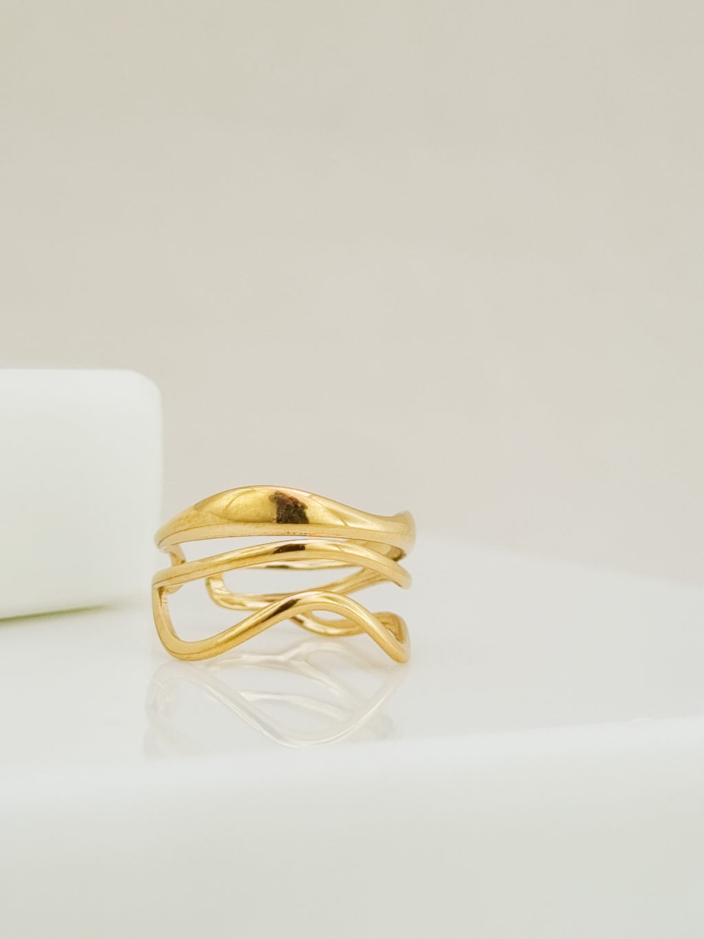 A gold-plated abstract wrap ring with a fluid, organic wavy design, displayed on a clean white surface with a soft reflection.