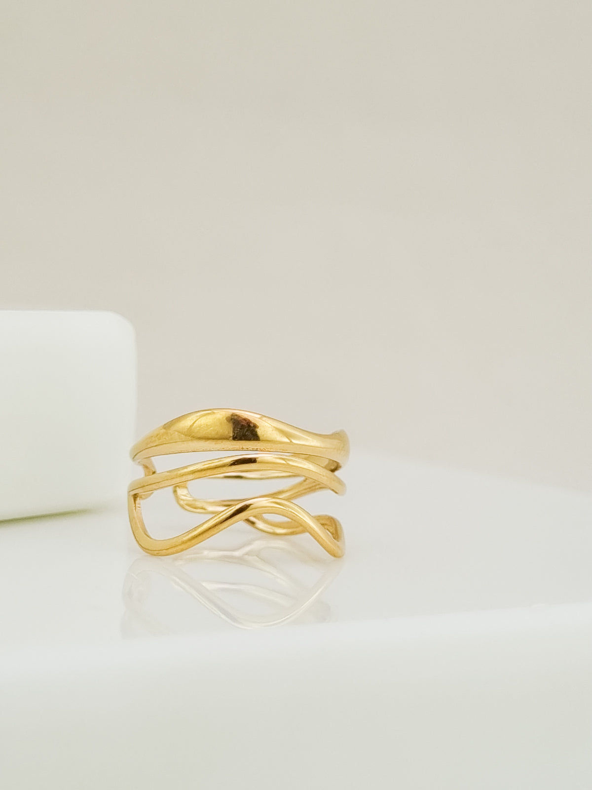 A gold-plated abstract wrap ring with a fluid, organic wavy design, displayed on a clean white surface with a soft reflection.