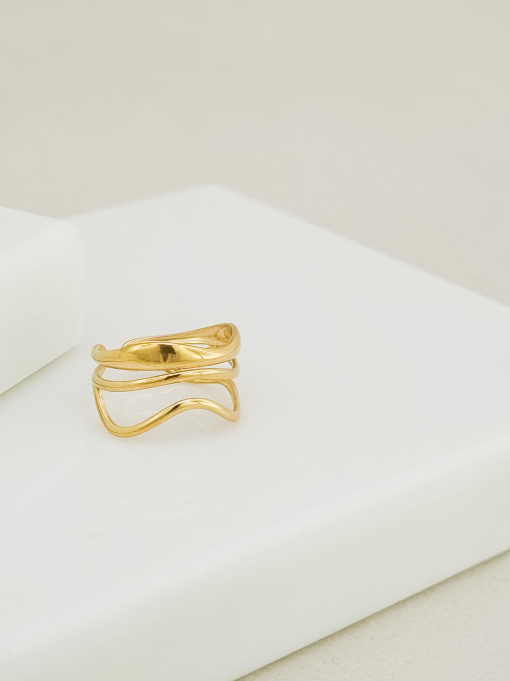 A gold wavy open ring with three parallel curved bands, displayed on a clean white geometric block against a light beige background.