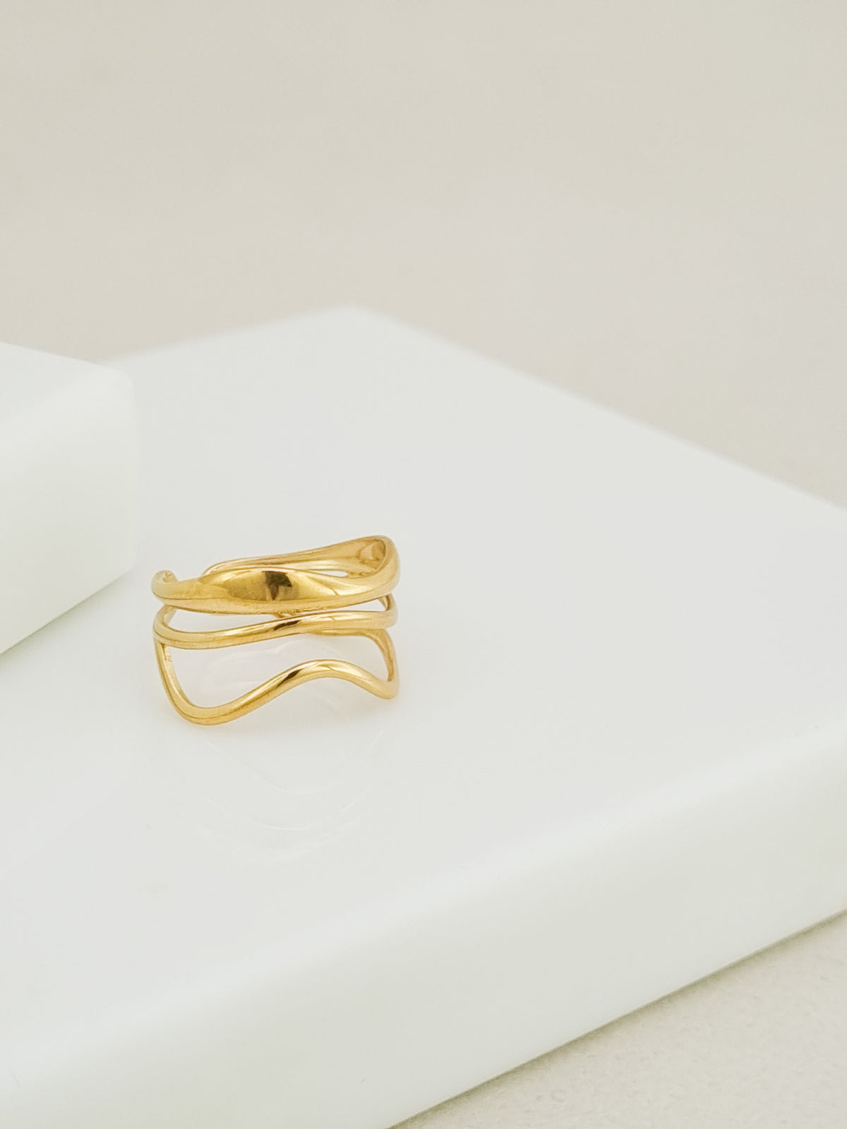 A gold wavy open ring with three parallel curved bands, displayed on a clean white geometric block against a light beige background.