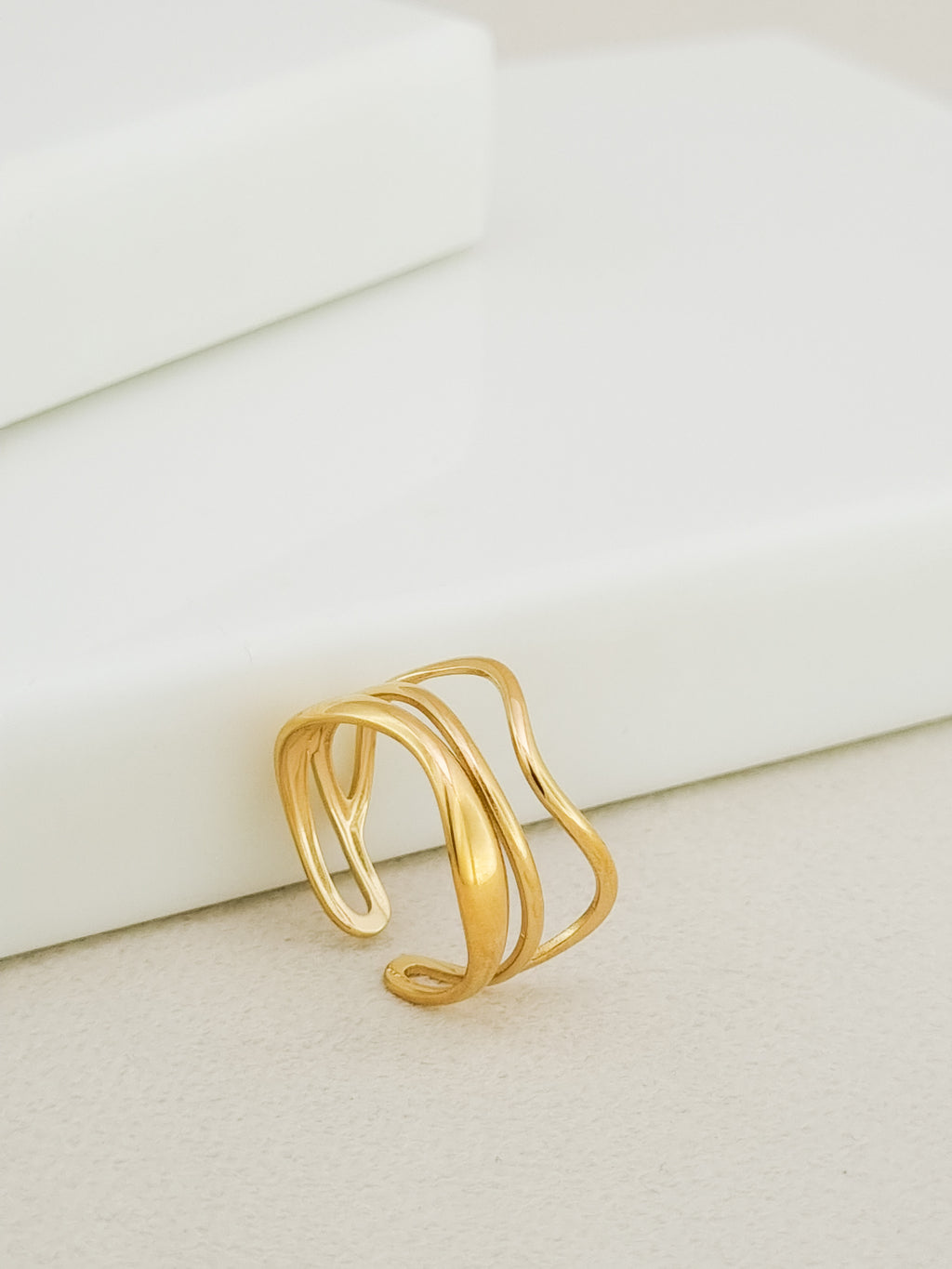 A gold-plated multi-band ring with a wavy, fluid design, resting on a light beige textured surface next to a white geometric block.