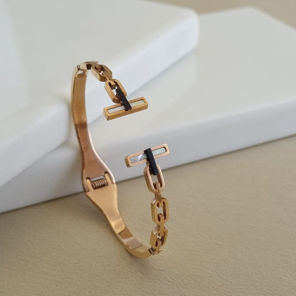 An 18K rose gold-plated open-ended cuff bangle featuring a chain-link design and T-bar terminals with white inlays, displayed on a neutral surface.