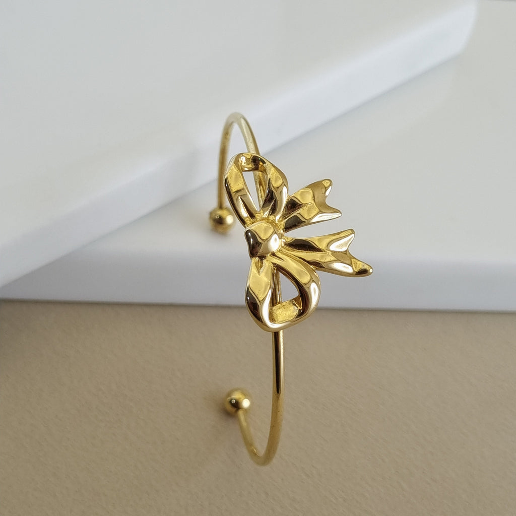 A high-shine 18K gold-plated open cuff bangle featuring a central sculpted ribbon bow, shown resting on a minimalist white and beige surface.