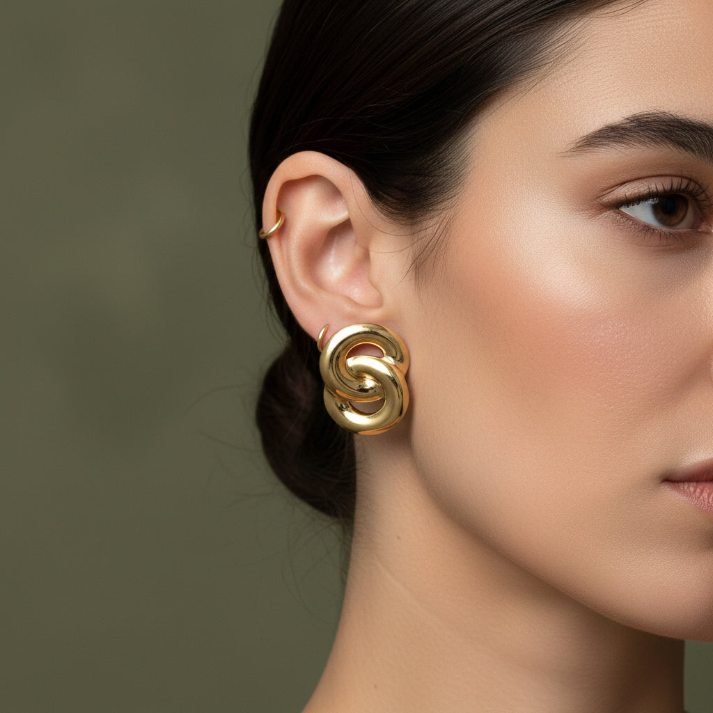 A woman with a sleek bun wearing large, 18K gold-plated interlocked knot stud earrings with a high-polish finish.