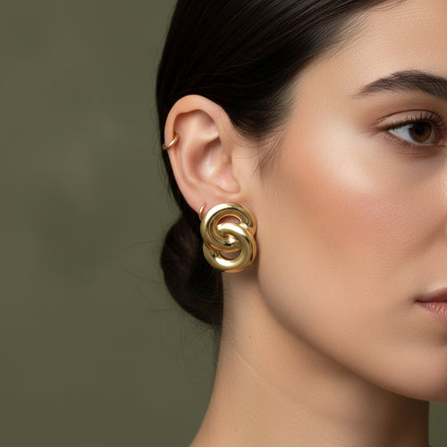 A woman with a sleek bun wearing large, 18K gold-plated interlocked knot stud earrings with a high-polish finish.