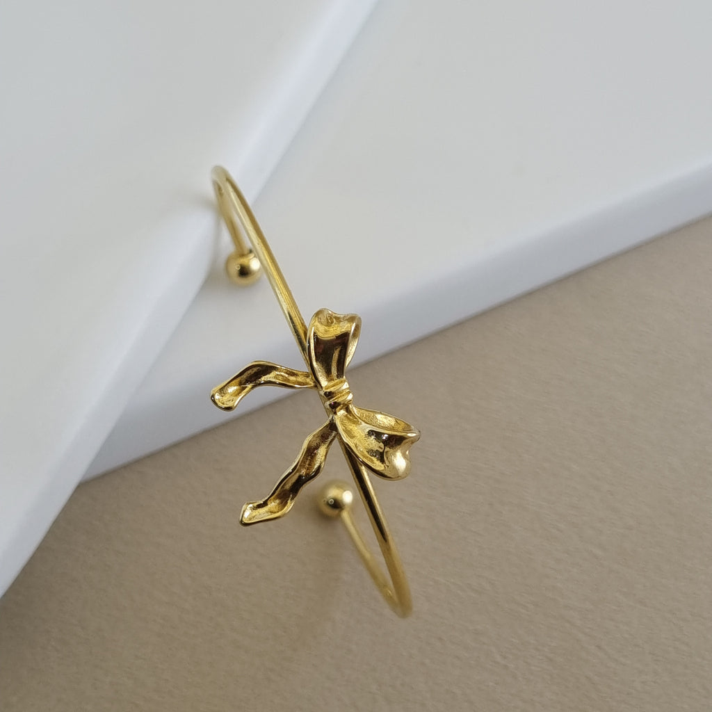 A high-polish 18K gold-plated organic wave bangle with a molten texture, shown resting on a minimalist white and beige background.