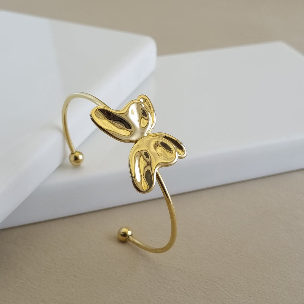 A high-shine 18K gold-plated open cuff bangle featuring a central sculpted butterfly or petal motif, displayed on a minimalist white surface.