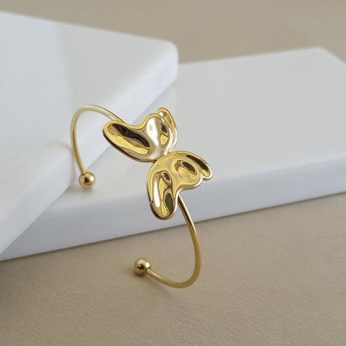 A high-shine 18K gold-plated open cuff bangle featuring a central sculpted butterfly or petal motif, displayed on a minimalist white surface.