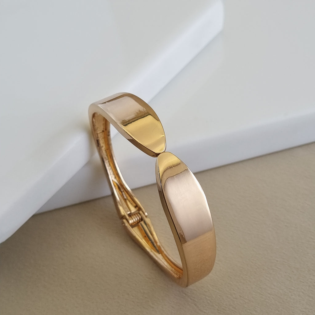 A high-polish 18K rose gold-plated hinged bangle with a tapered, sculptural geometric design, displayed on a minimalist white and beige surface.