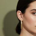 A woman with a sleek ponytail wearing large, 18K gold-plated sculptural drop earrings in a sharp, tapered prism design.