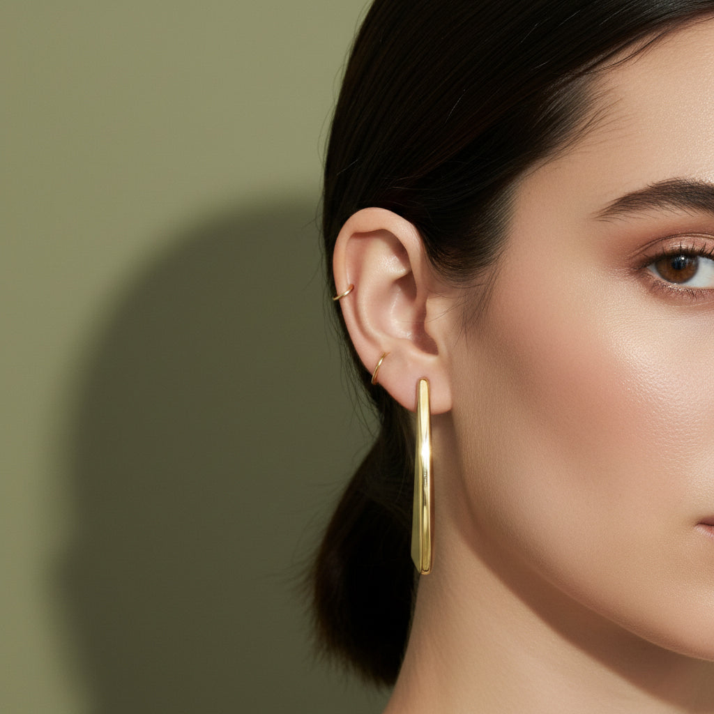A woman with a sleek ponytail wearing large, 18K gold-plated sculptural drop earrings in a sharp, tapered prism design.
