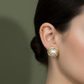 A woman with a sleek bun wearing large, 18K gold-plated stud earrings featuring a central pearl surrounded by a high-polish gold spiral.