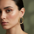 A woman with a sleek hairstyle wearing large, 18K gold-plated vertical oval hoop earrings with a high-polish tubular design.