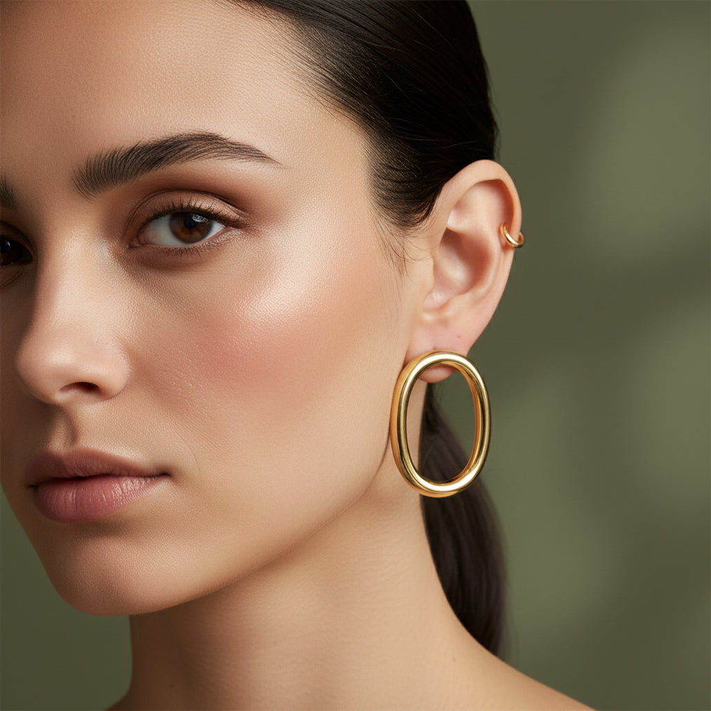 A woman with a sleek hairstyle wearing large, 18K gold-plated vertical oval hoop earrings with a high-polish tubular design.