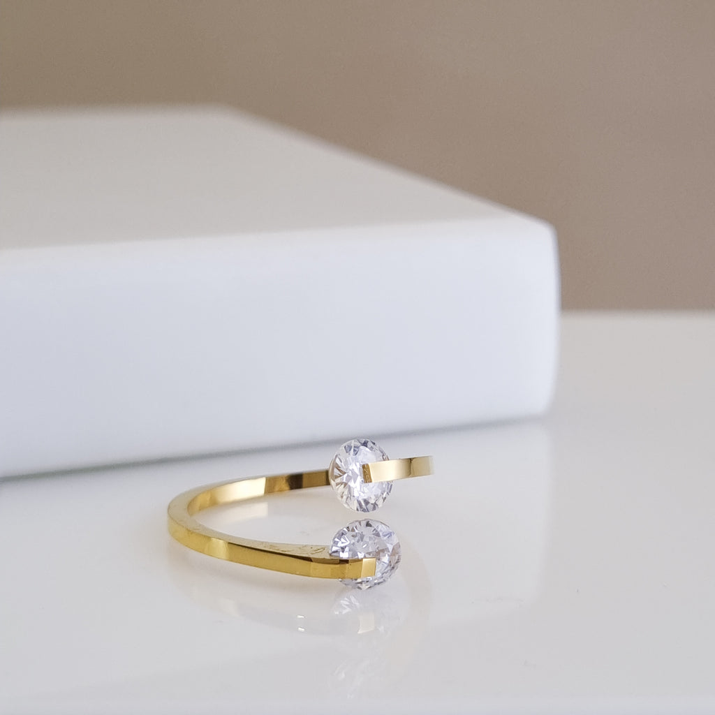 A high-polish 18K gold-plated, size-adjustable open-ended ring featuring two brilliant-cut crystals in a bypass setting, displayed on a minimalist white surface.