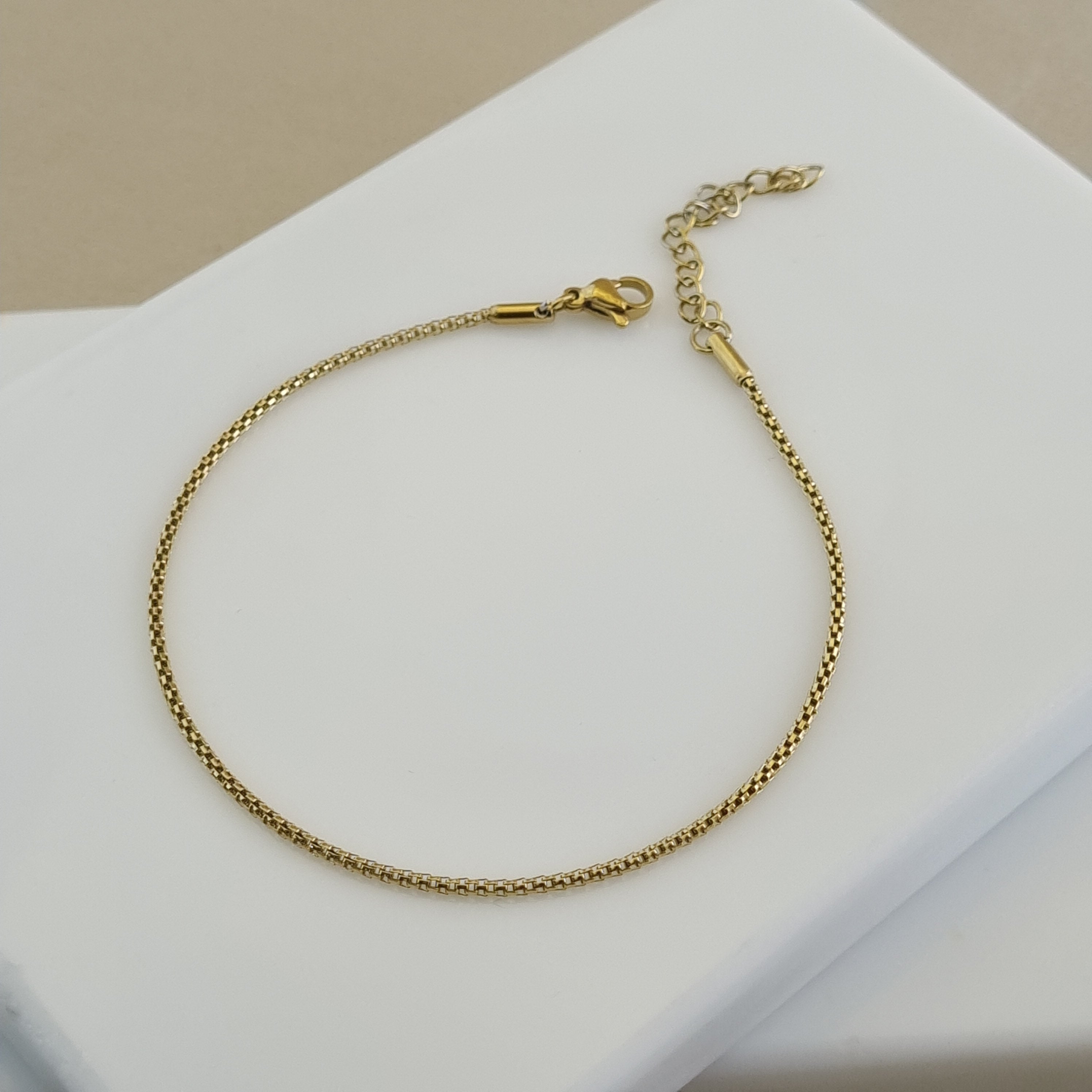 Close-up of an 18K gold plated box chain anklet featuring a smooth geometric design and an adjustable lobster clasp.