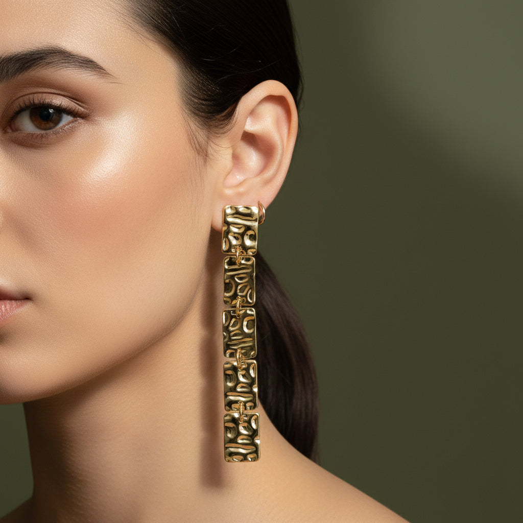 A woman with a low ponytail wearing long, 18K gold-plated drop earrings consisting of five linked rectangular segments with a hammered, molten texture.