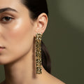 A woman with a low ponytail wearing long, 18K gold-plated drop earrings consisting of five linked rectangular segments with a hammered, molten texture.