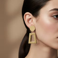 A woman with a sleek low ponytail wearing 18K gold-plated drop earrings featuring a circular top and a large, hollow trapezoid-shaped dangle.