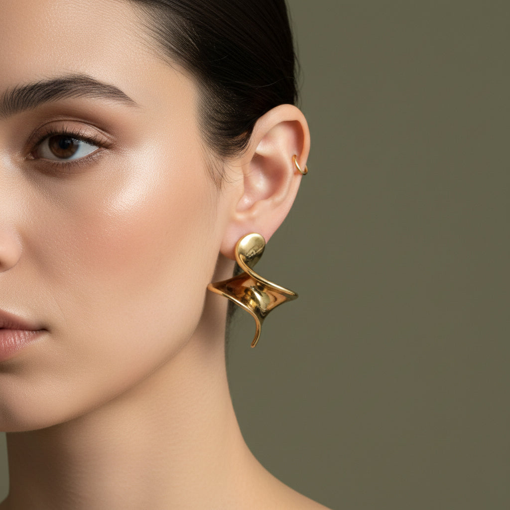 A woman with a sleek hairstyle wearing large, 18K gold-plated sculptural twist hoop earrings with a high-polish fluid design.