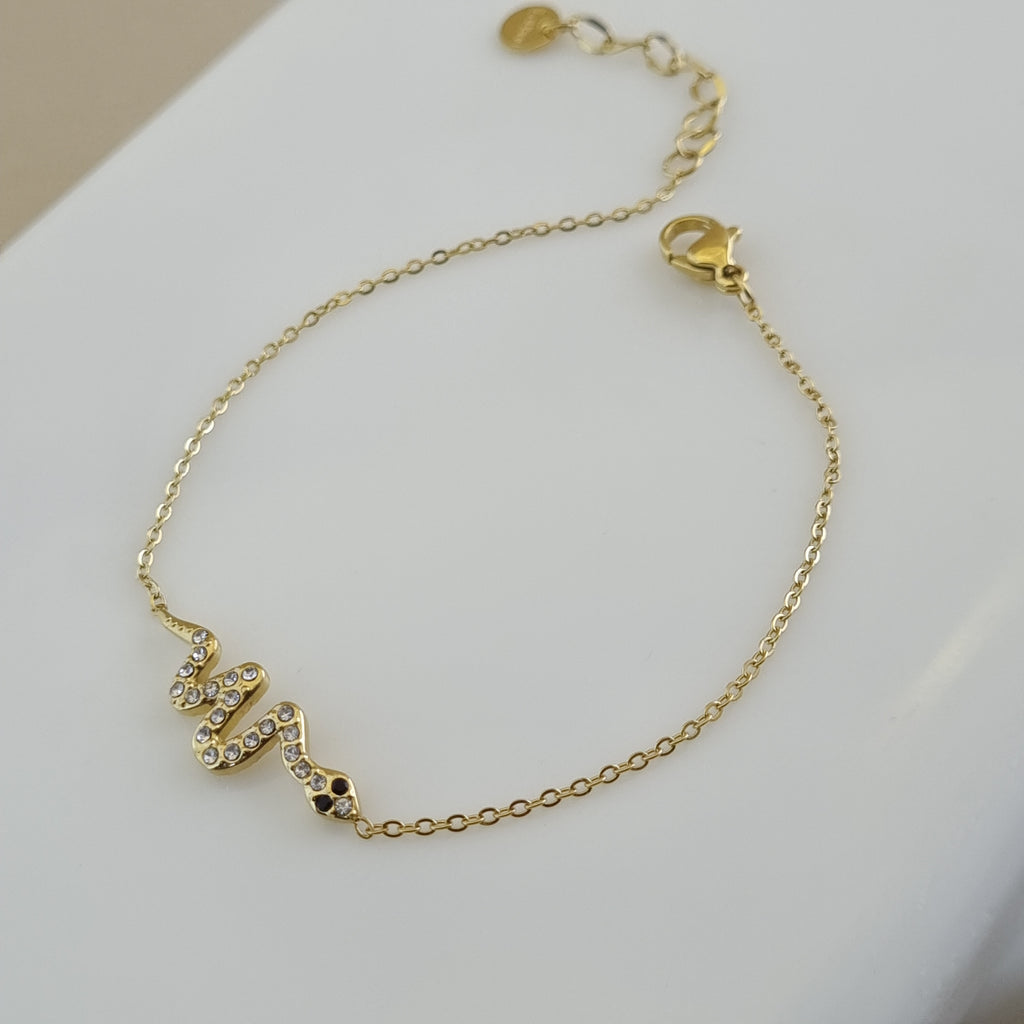Treshire 18K gold plated serpent bracelet with pave stones and dark eyes on a gold rope chain, displayed on a white marble slab.