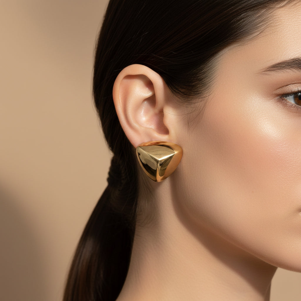 A woman with a sleek ponytail wearing large, 18K gold-plated triangular prism stud earrings with a high-polish reflective finish.