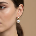 A woman with a soft brown ponytail wearing 18K gold-plated rectangular huggie earrings with a large suspended white pearl drop.