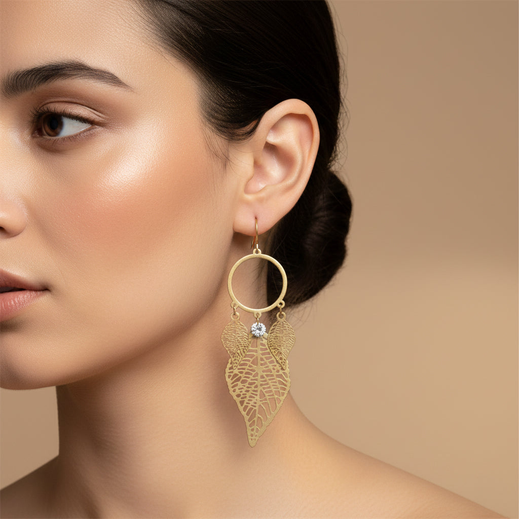 A woman with a sleek low bun wearing large, 18K gold-plated filigree leaf drop earrings featuring a circular hoop and three cascading golden leaves.