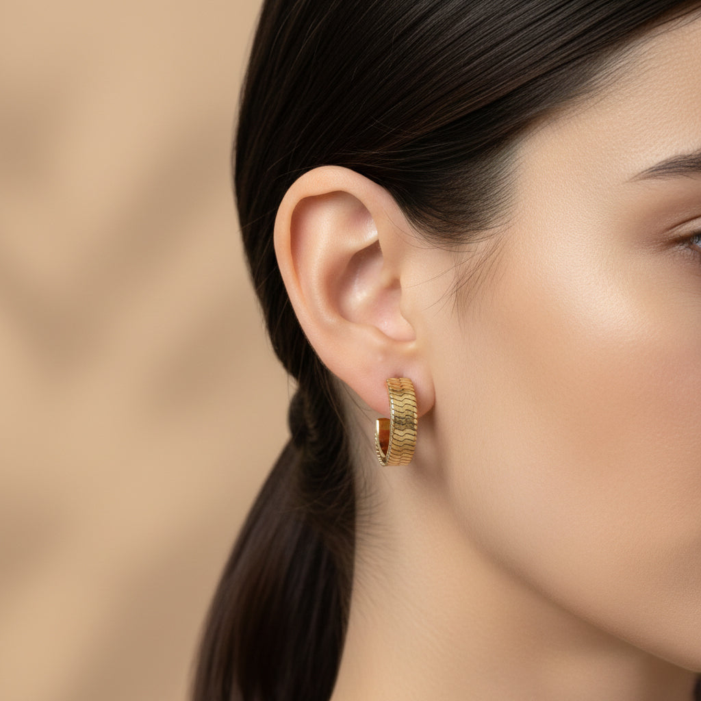 A woman with a sleek ponytail wearing 18K gold-plated rectangular huggie earrings featuring a repeating embossed chevron pattern and a high-polish finish.