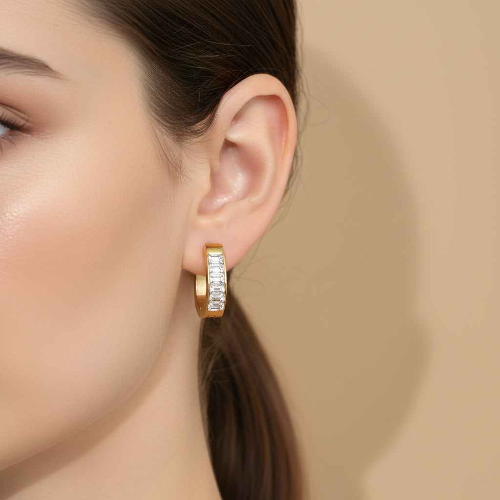 A woman with a sleek ponytail wearing 18K gold-plated rectangular huggie earrings featuring a row of clear baguette-cut crystals and a high-polish finish.