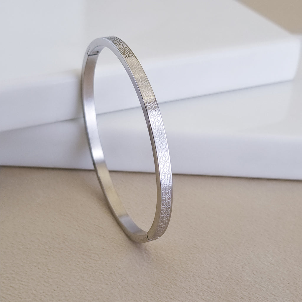 A high-polish silver-toned hinged bangle featuring a laser-engraved honeycomb pattern, displayed on a minimalist white and beige background.