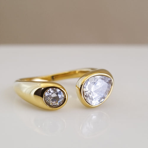 A high-polish 18K gold-plated open-ended ring featuring two differently sized brilliant-cut crystals, displayed on a minimalist white surface.